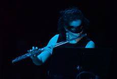 wearing a mask over her eyes and cast in dark blue light, flutist Sarah Frisoff performs in front of a black background.